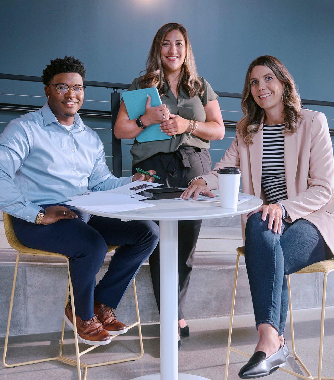 Two female employees and one male employee smiling and gathered around a tall white table.