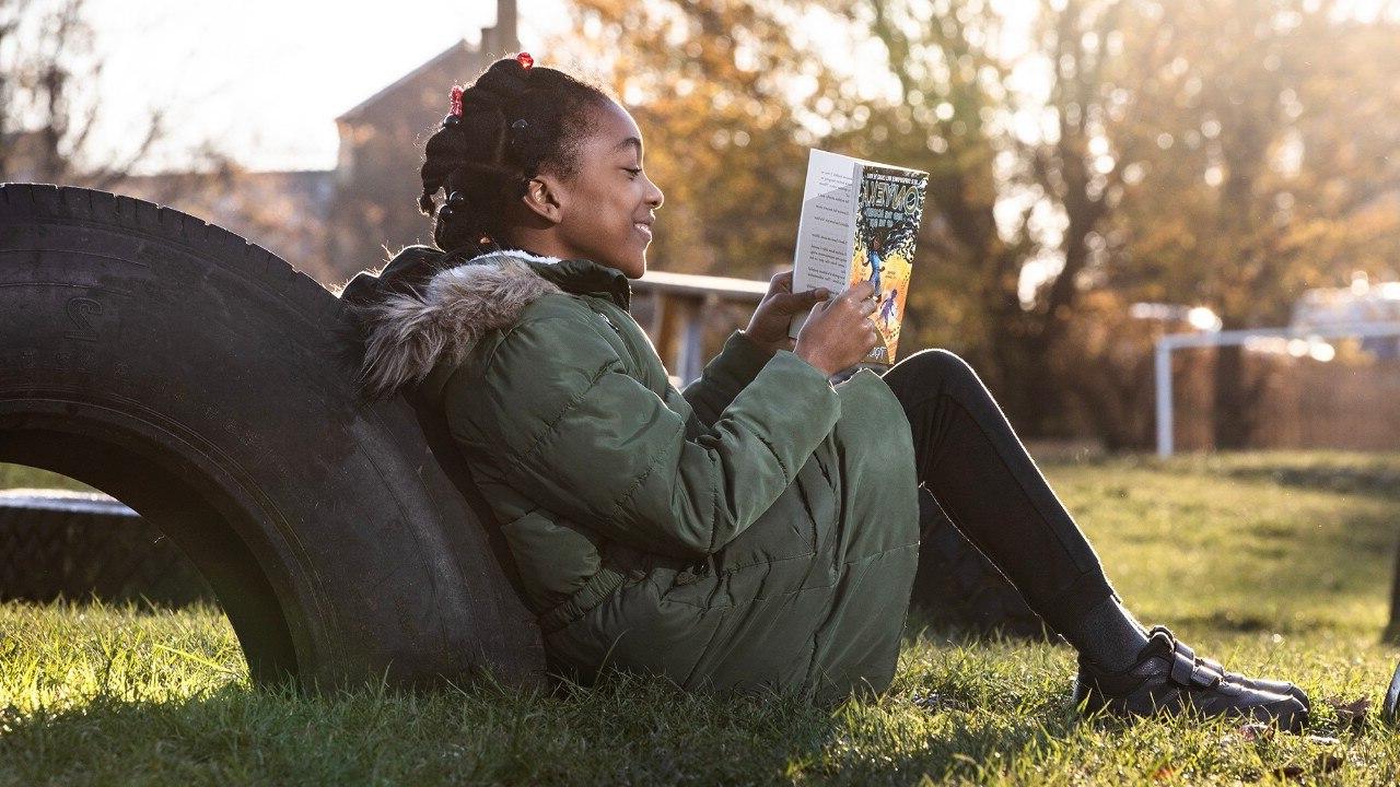 一位年轻的女士，坐在绿草地上，倚在一个大轮胎上看书.