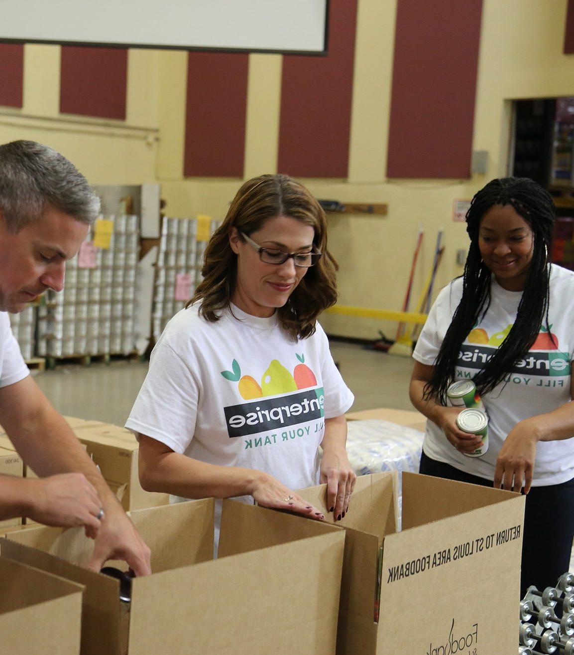 Two female Enterprise employees and one male employee, all wearing white Enterprise Fill Your Tank t-shirts, pack boxes during a community service project.