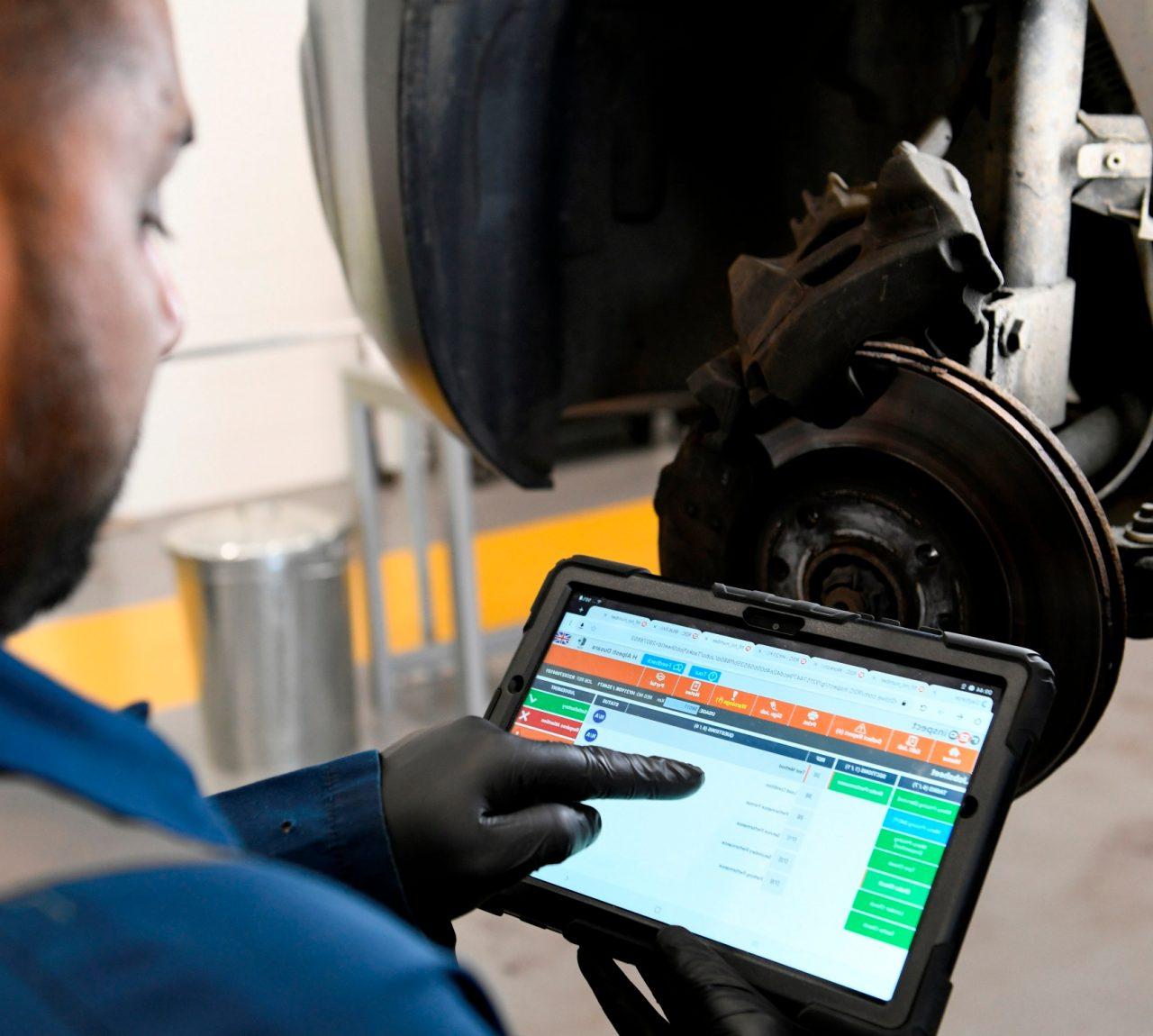 A male Enterprise employee, wearing a black glove in an auto mechanic's garage, points at an iPad.